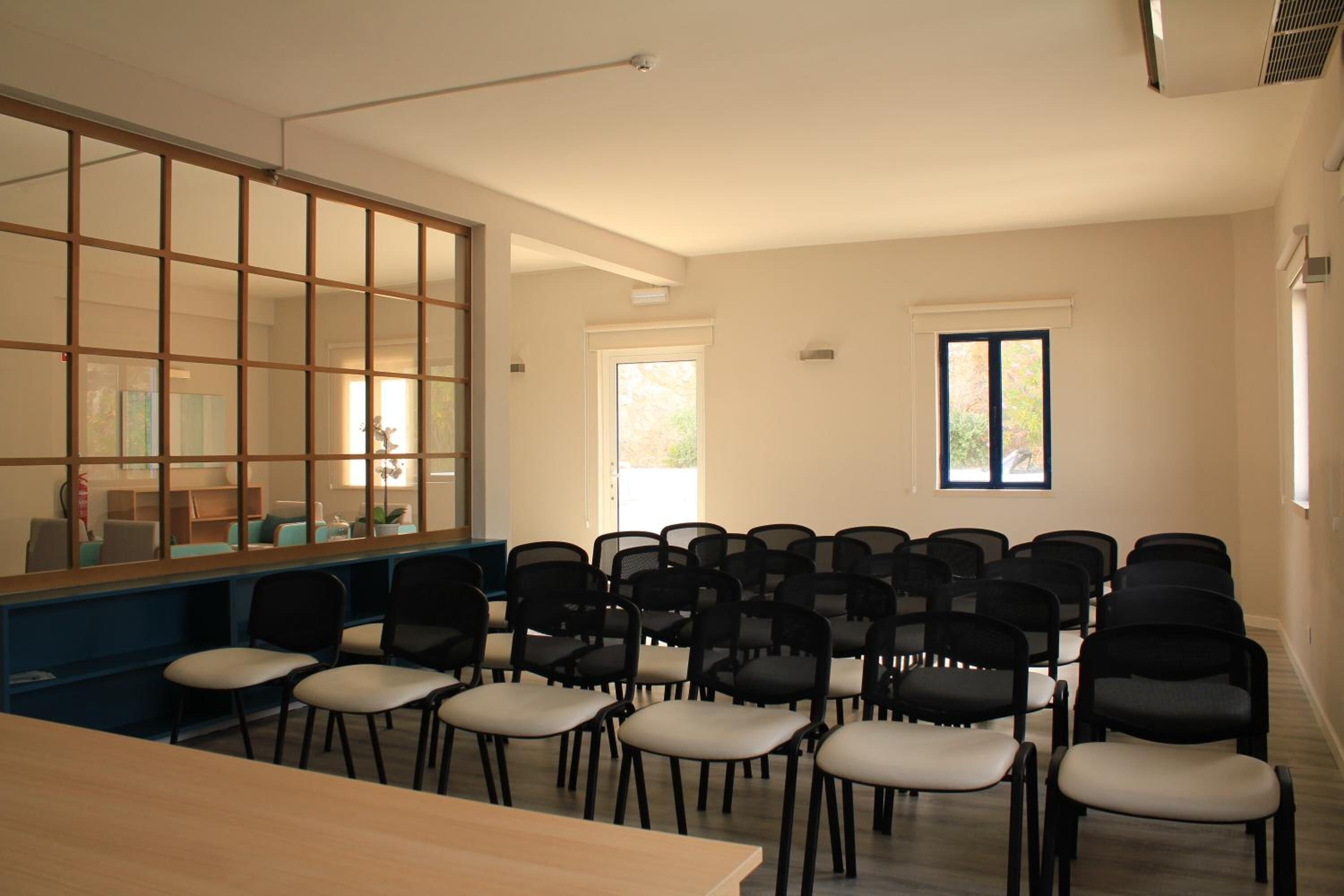 A conference room with a long table and rows of chairs, large windows, and a view of the outdoors.