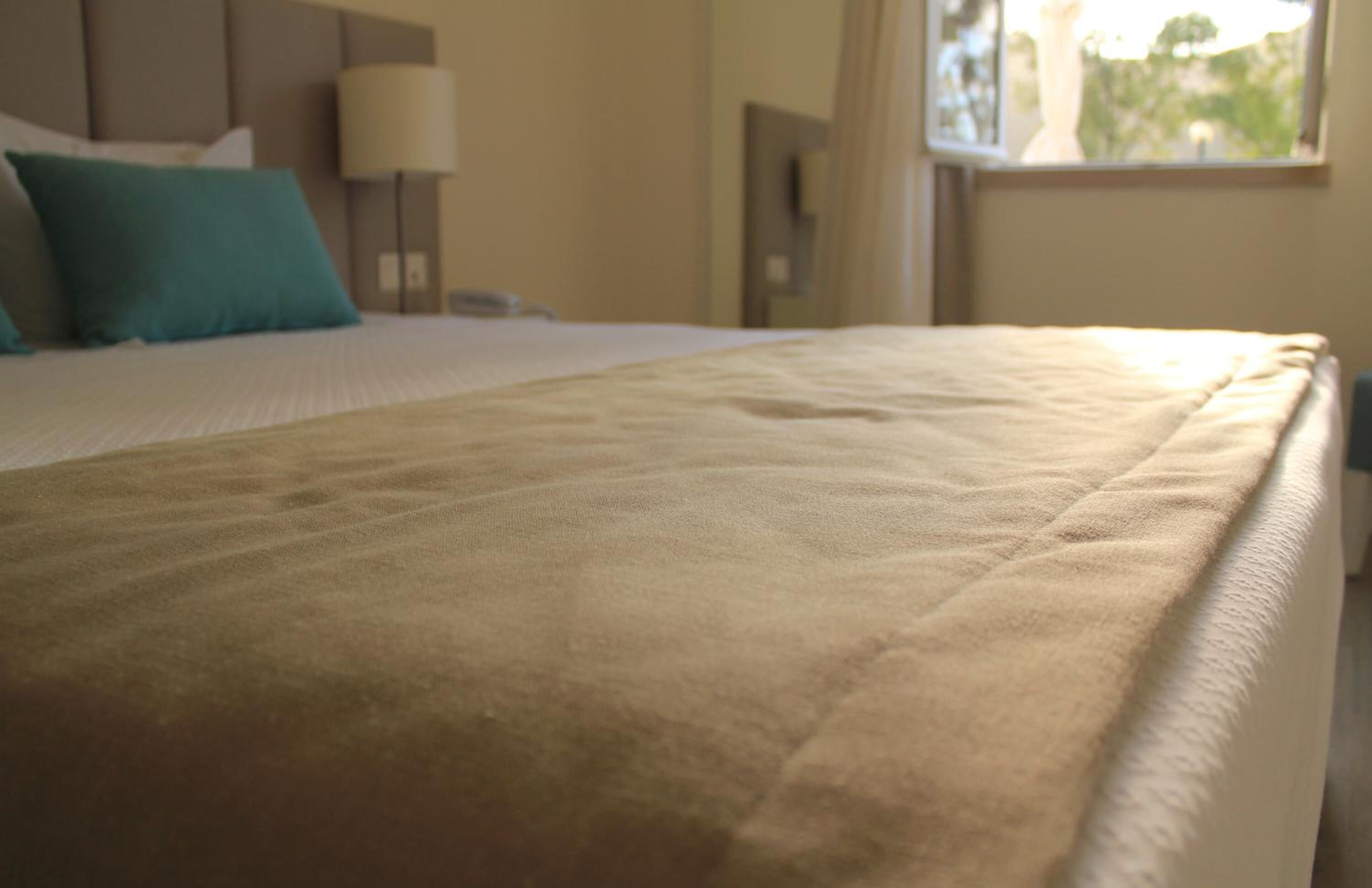 Una cama bien tendida con una manta color beige, una almohada verde aguamarina y una lámpara en una mesita de noche.