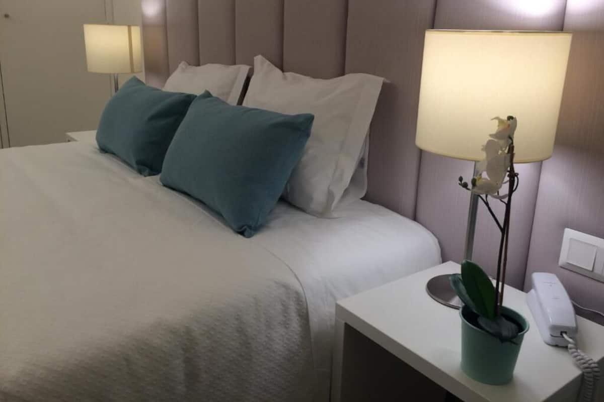 Een net opgemaakt bed met witte en teal kussens, een bedlampje, een kleine potplant en een telefoon op het nachtkastje.