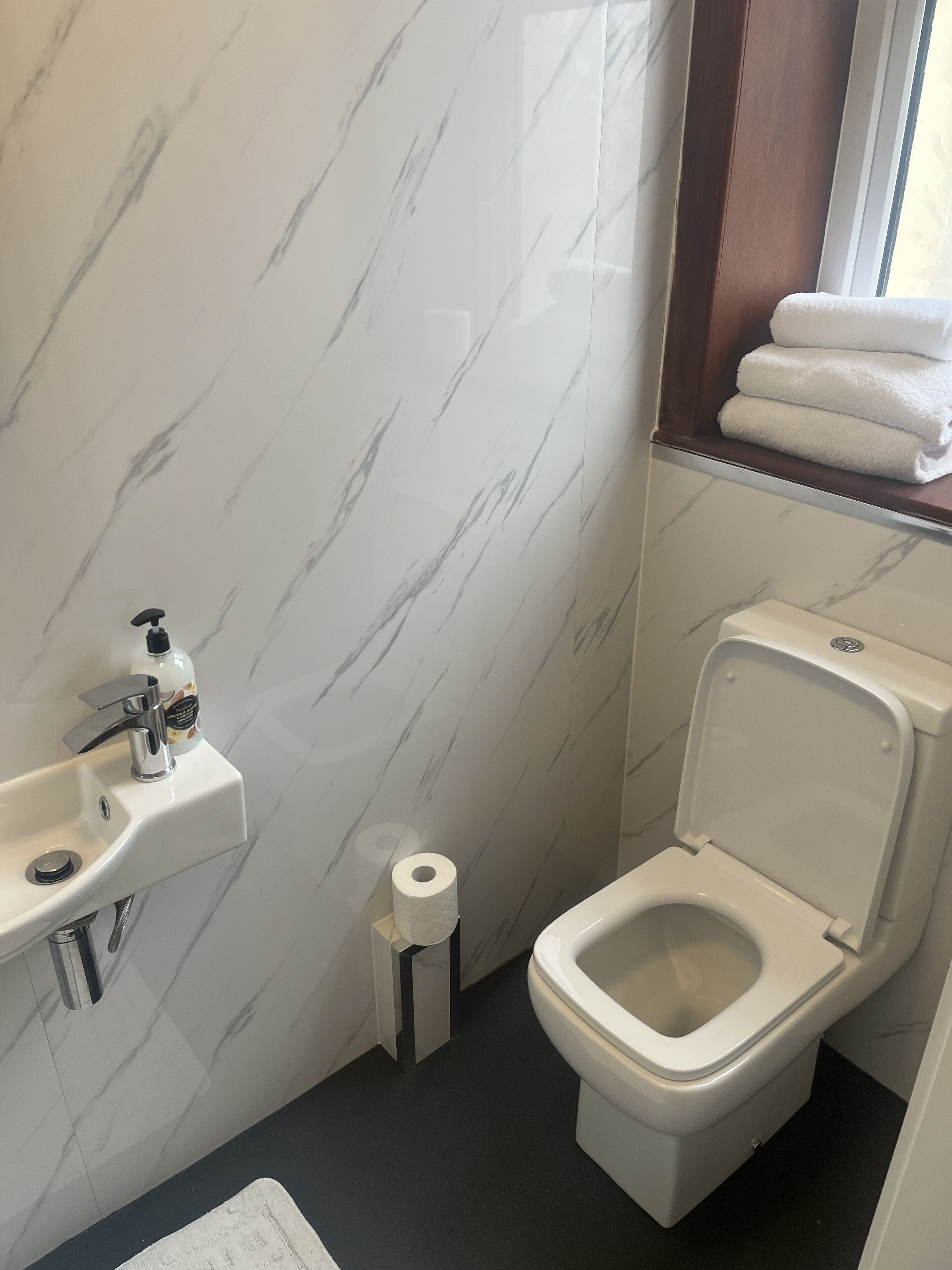 A bathroom with a marble wall, a sink with a soap dispenser, a toilet, and a window with towels.