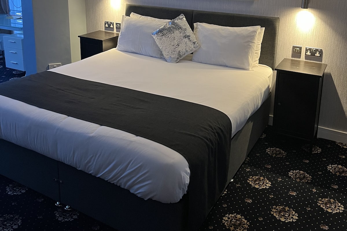 Ein Hotelzimmer mit einem großen Bett, zwei Nachttischen mit Lampen, einem an der Wand hängenden Stadtbild und einem gemusterten Teppich.