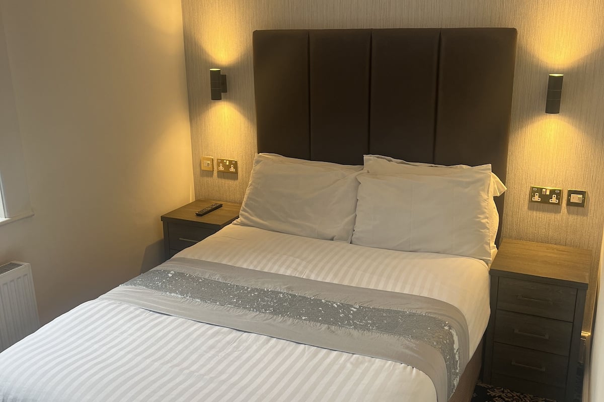 Ein ordentlich bezogenes Bett mit weißen Laken, einem dunklen Kopfteil und zwei Nachttischlampen.