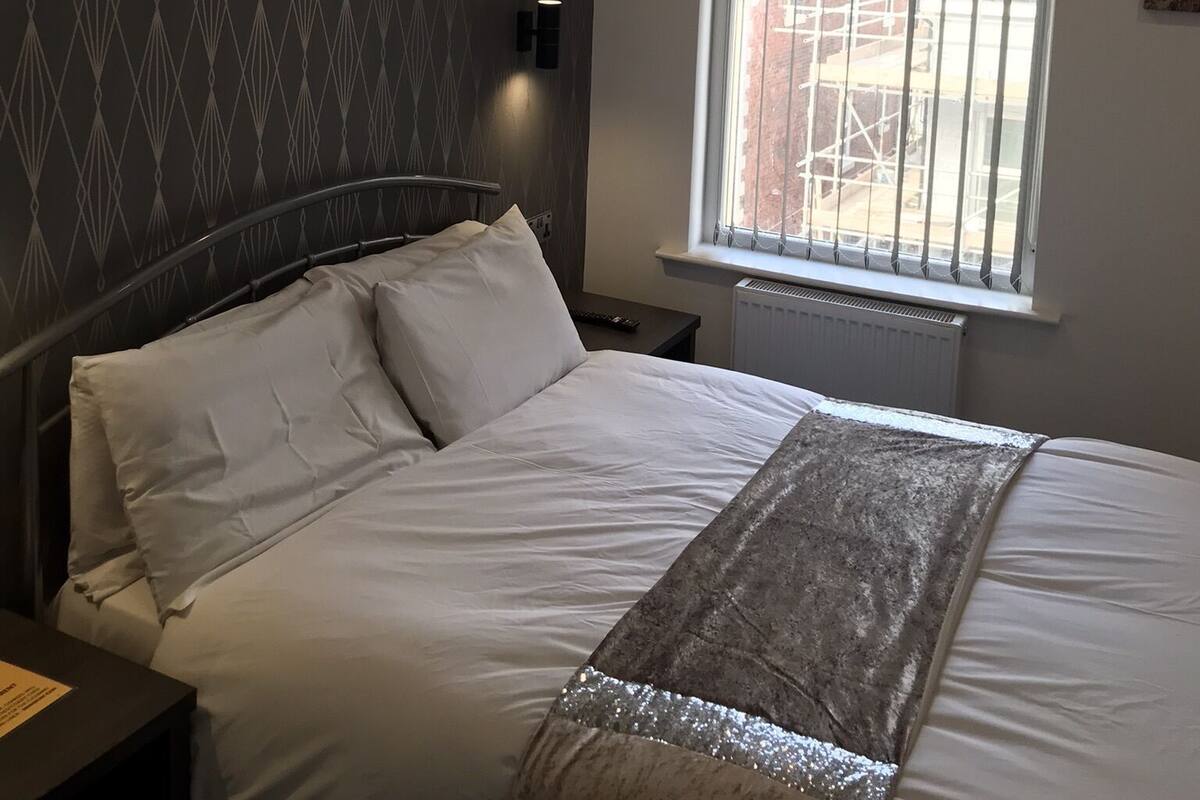 Ein Schlafzimmer mit einem Bett, einem Nachttisch, einer Wandleuchte, einem Fenster mit Jalousien und Blick nach außen.