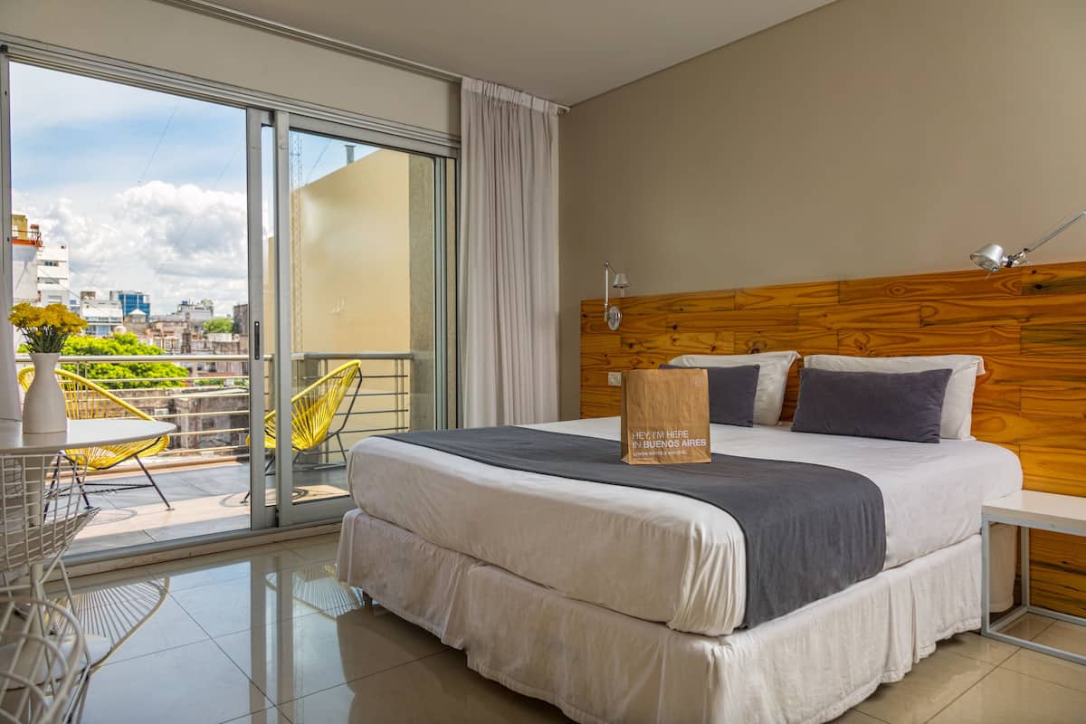 A hotel room with a large bed, a wooden headboard, a balcony with yellow chairs, and a view of the city.