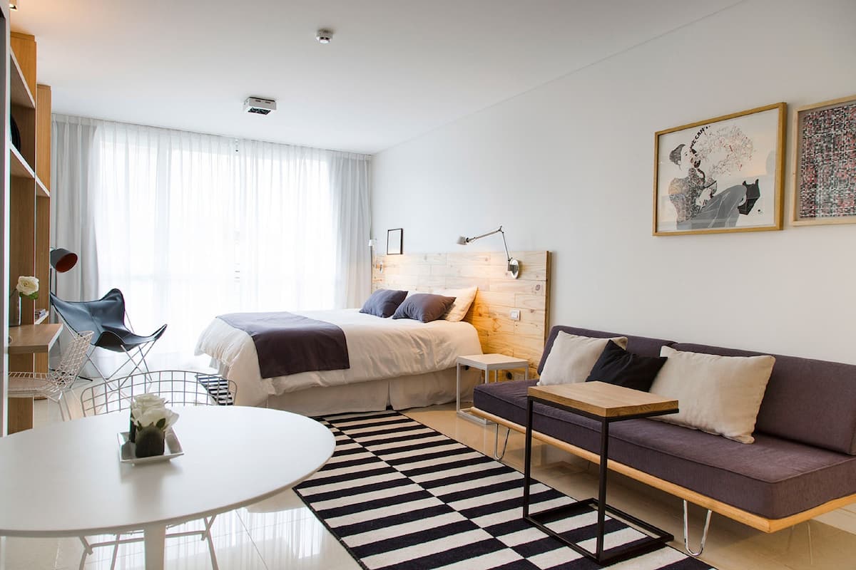 A modern hotel room with a bed, a sofa, a dining table, and a chair. There are two paintings on the wall and a window with curtains.
