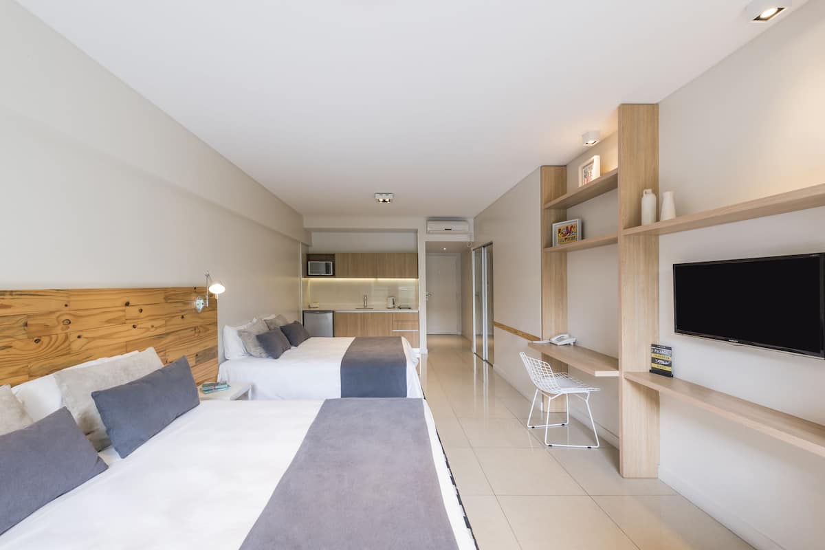 A modern hotel room with two beds, a kitchenette, a TV, and a small seating area.