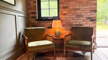 Une pièce avec deux fauteuils, une petite table avec une lampe, une fenêtre donnant sur un paysage verdoyant et un mur en briques.