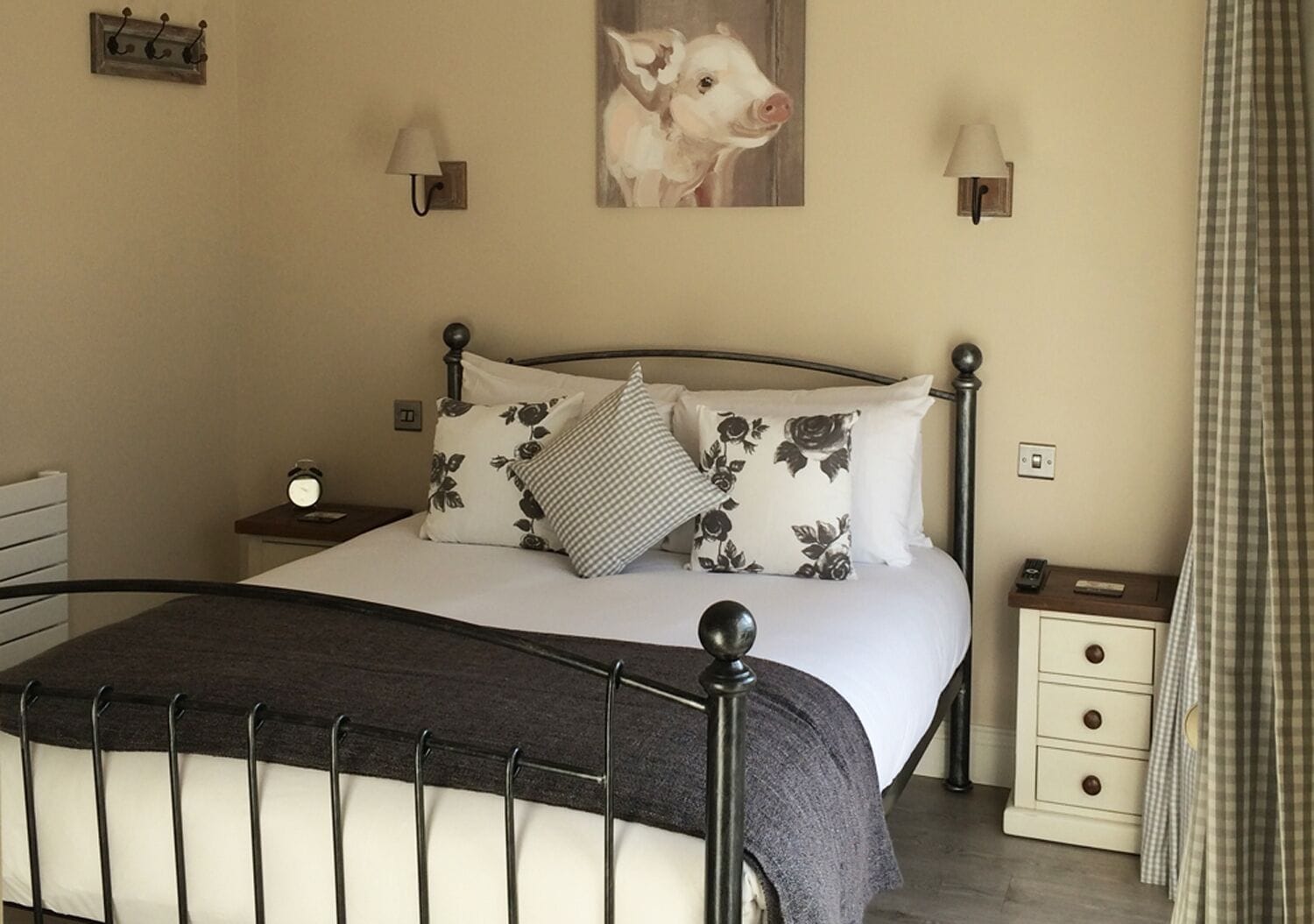 A neatly made bed with a metal frame, white bedding, and floral-patterned pillows. There are two bedside tables with lamps, a wall clock, and a painting of a cow.
