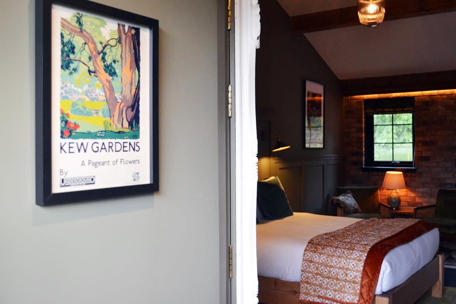 A framed artwork titled 'KEW GARDENS' by a Pageant of Flowers, a bed with a patterned blanket, a brick wall, and a window with a view of greenery.