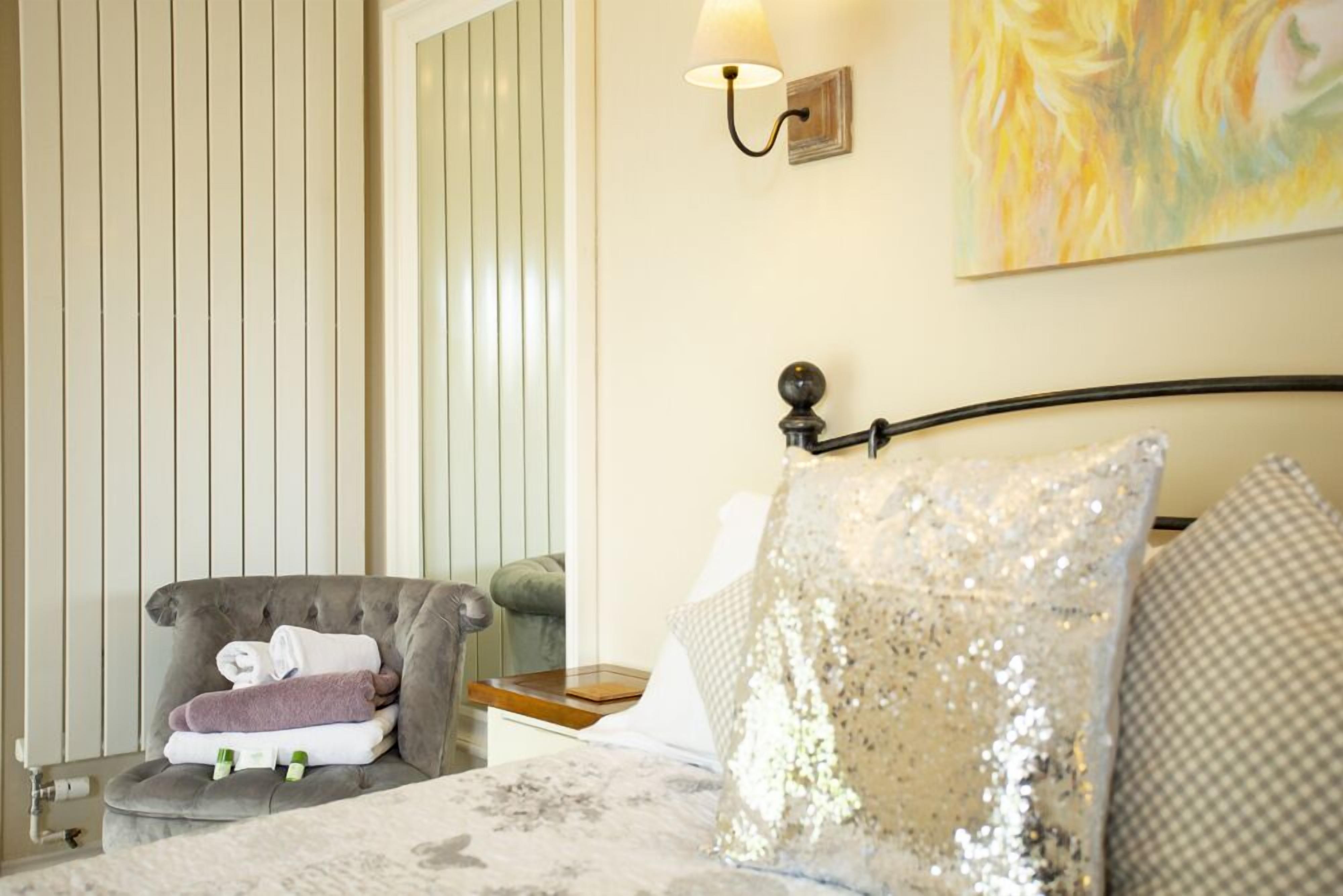A bedroom with a bed, a grey armchair with towels, a lamp, and a painting on the wall.