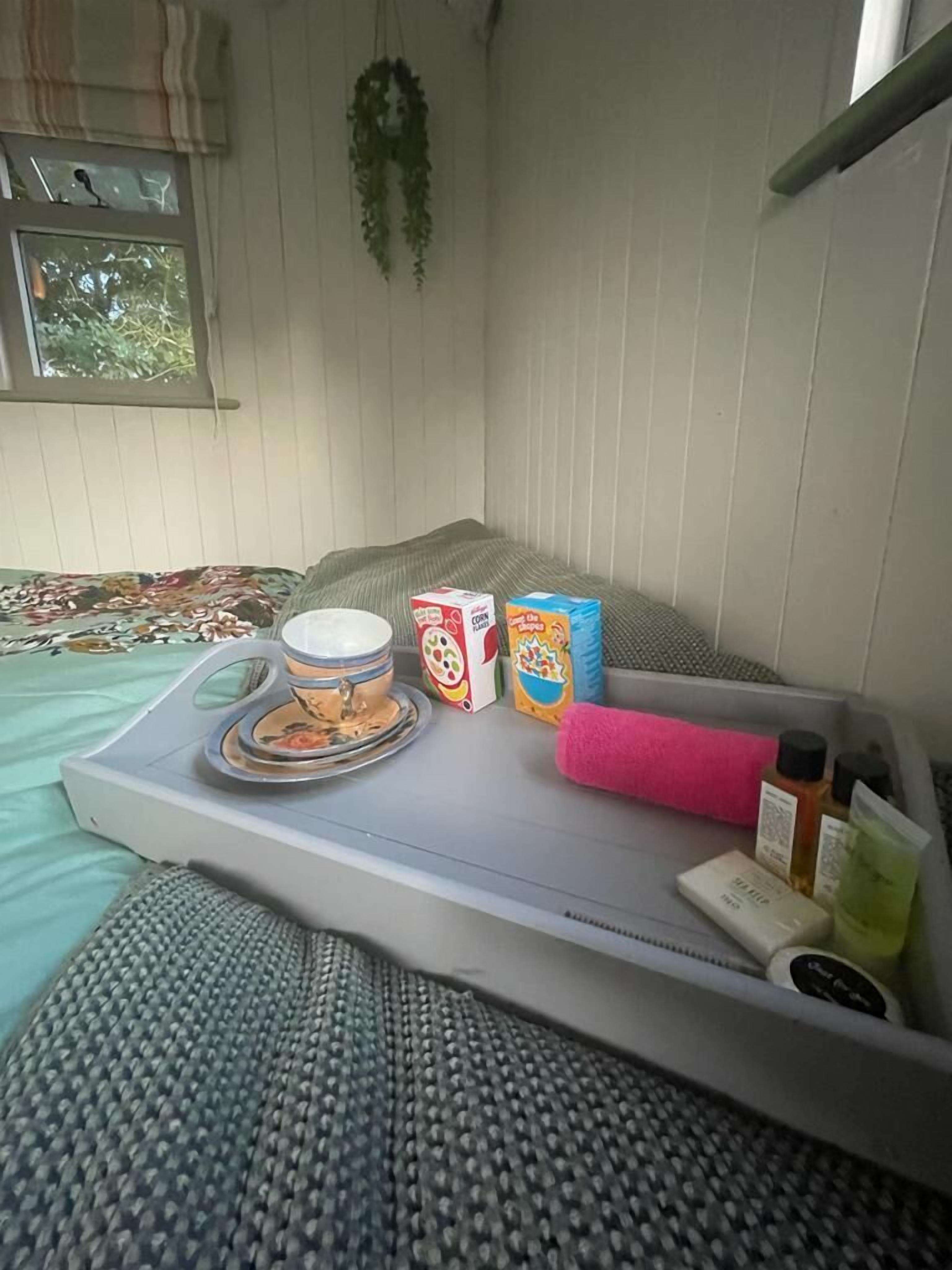A bed with a tray containing a teapot, cereal boxes, a pink towel, and toiletries.