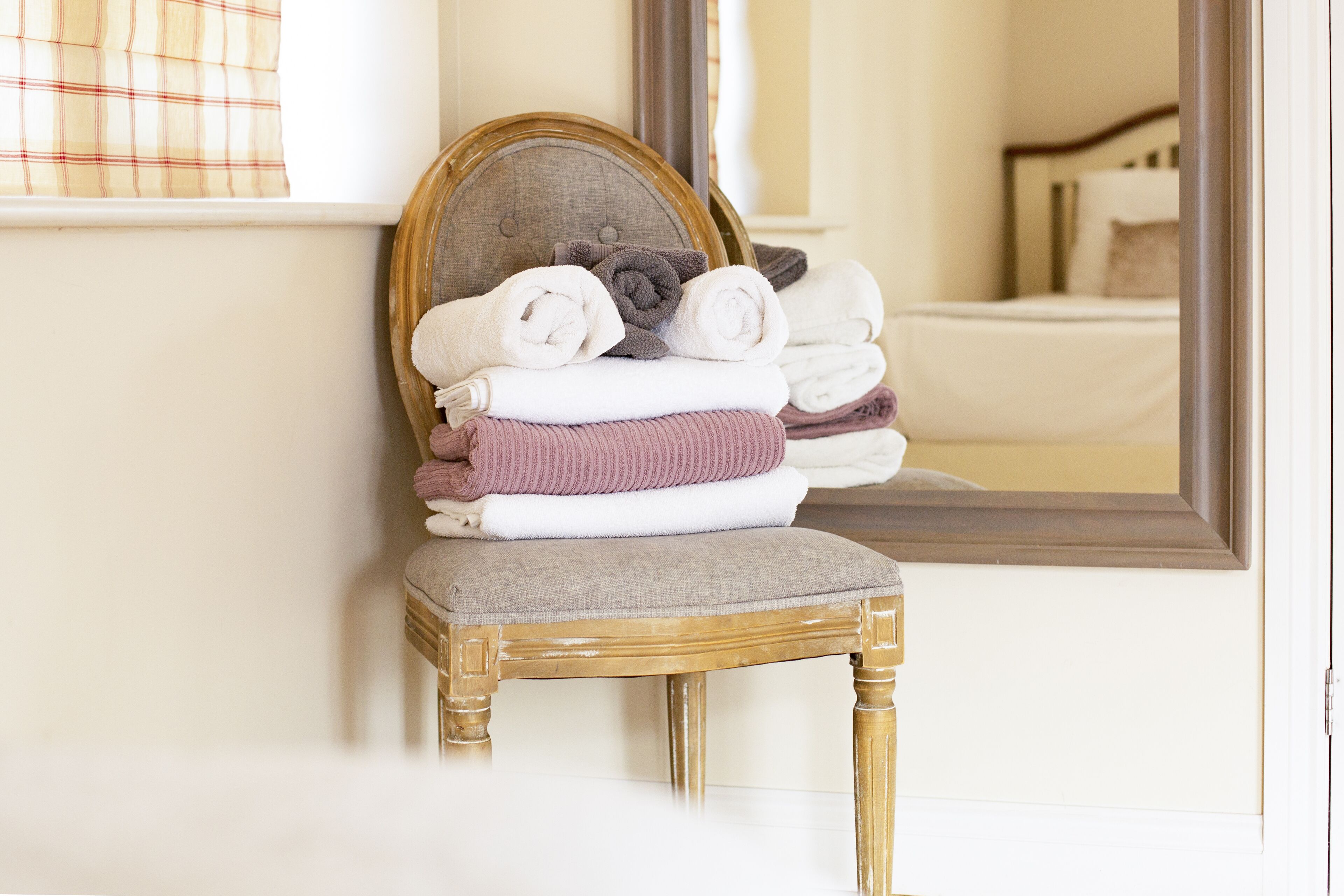 A stack of folded towels on a wooden chair in a room with a bed and a mirror reflecting the bed.