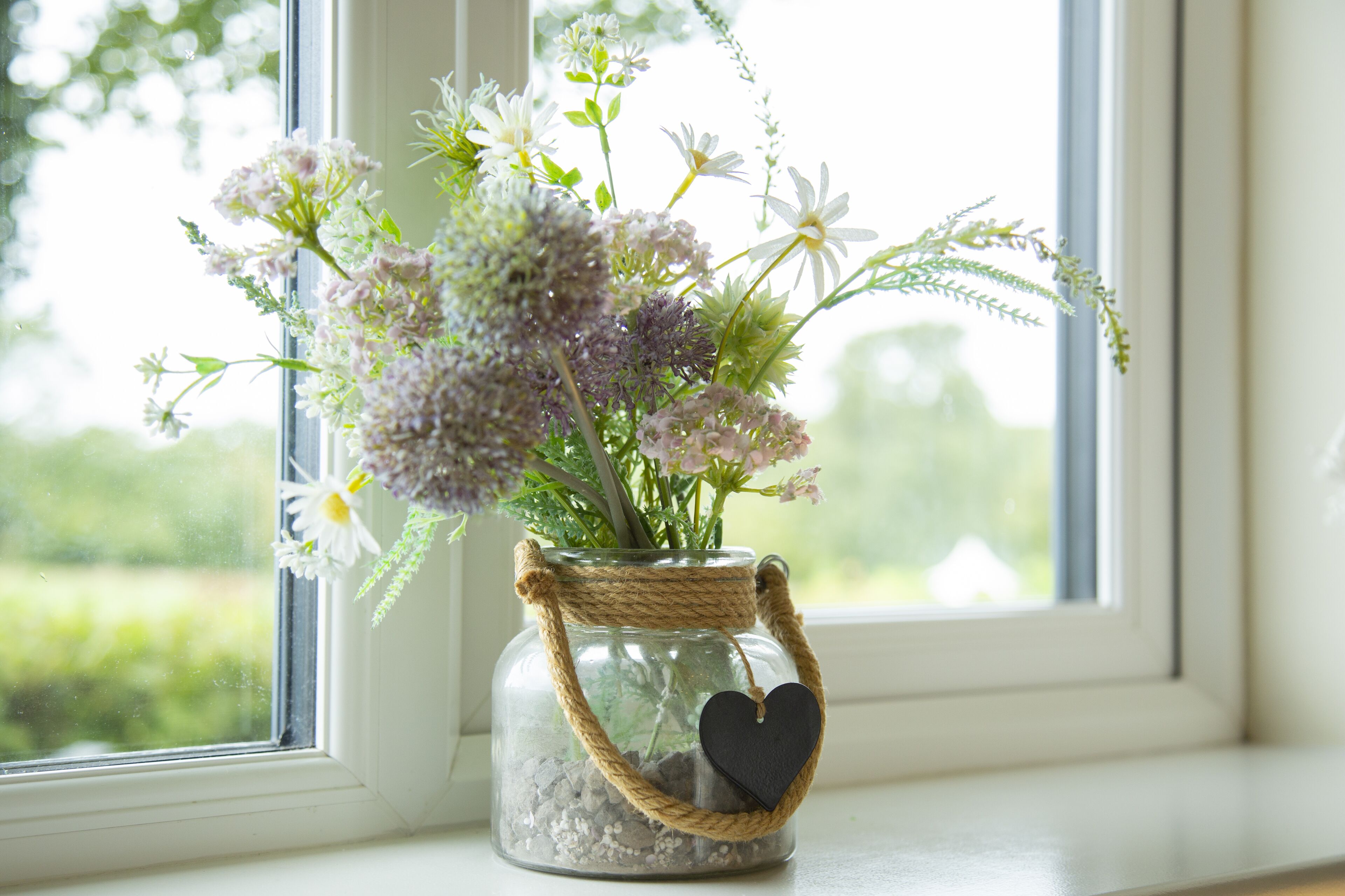 A glass jar with a rope handle and heart-shaped tag, containing flowers, placed on a windowsill.