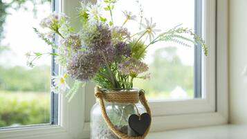 A glass jar with a rope handle and heart-shaped tag, containing flowers, placed on a windowsill.