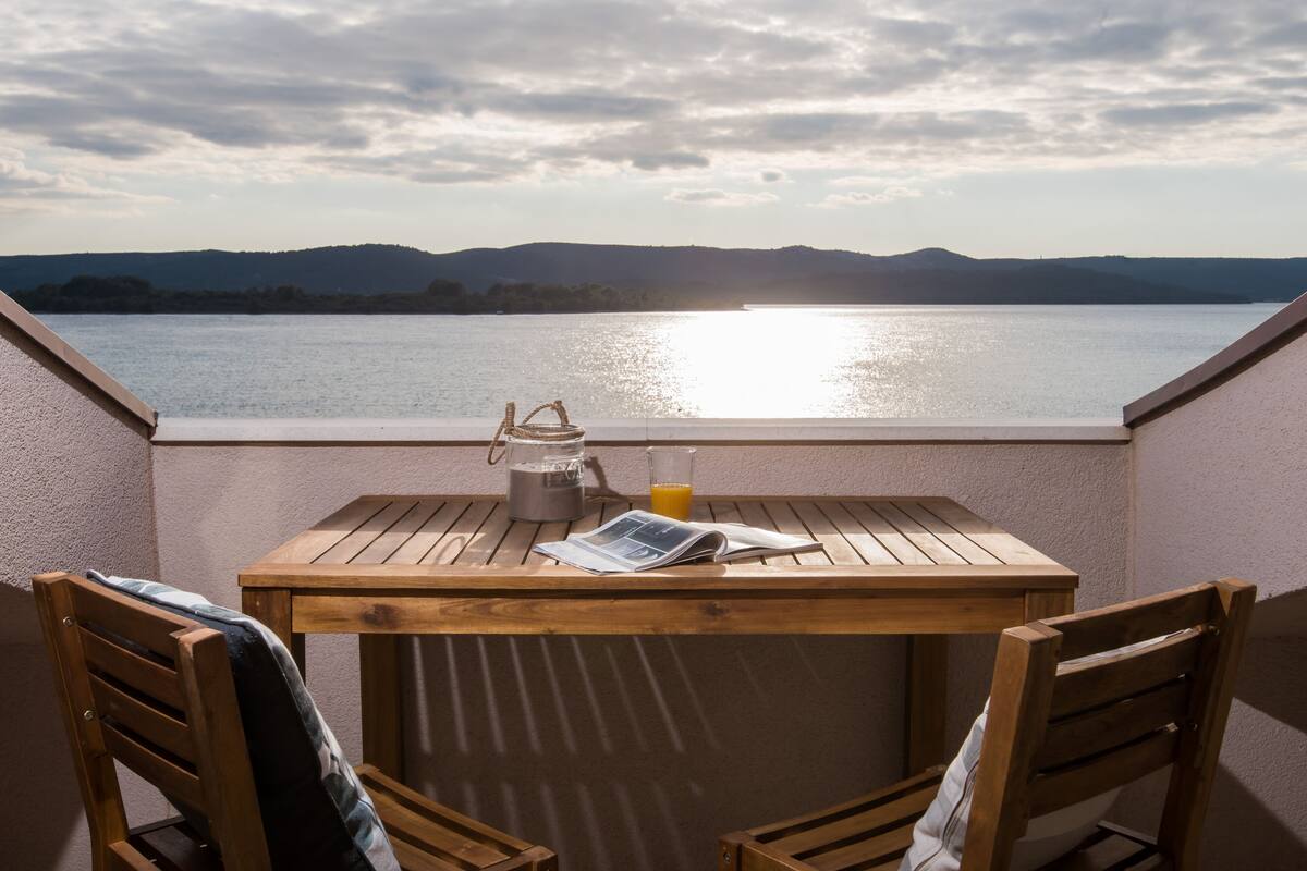 A wooden table with chairs on a balcony overlooking a body of water with mountains in the background.