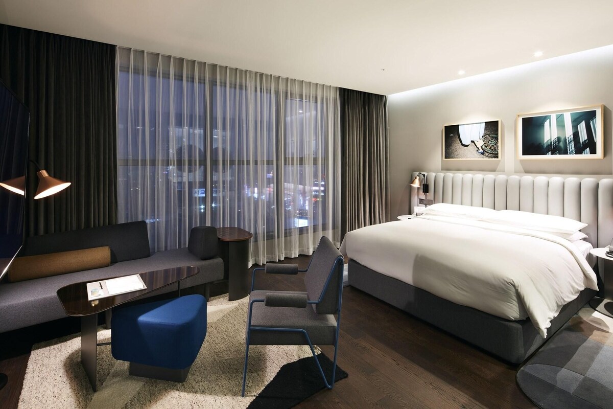 Ett modernt hotellrum med en stor säng, en soffa, en fåtölj, ett litet bord och utsikt över staden genom tunna gardiner.