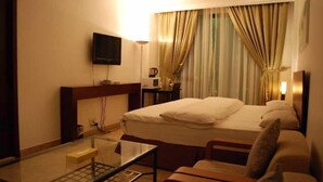 Quarto de hotel com cama, televisão, mesa de cabeceira, cadeira e janela com cortinas.