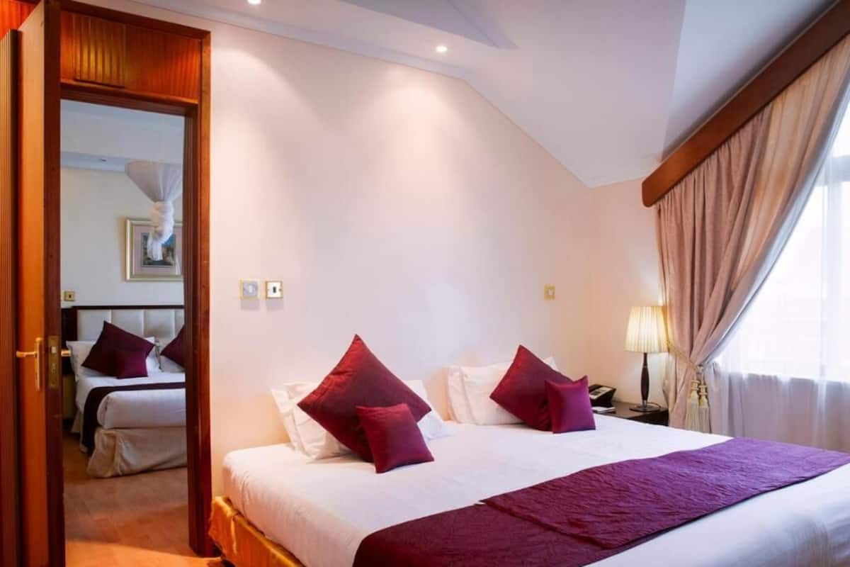 Una habitación de hotel con una cama grande, dos almohadas, una mesita de noche con lámpara y una ventana con cortinas.