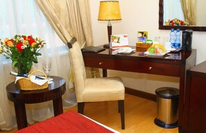 Habitación de hotel con escritorio, silla, mesita de noche con un florero, lámpara y un cesto de basura.