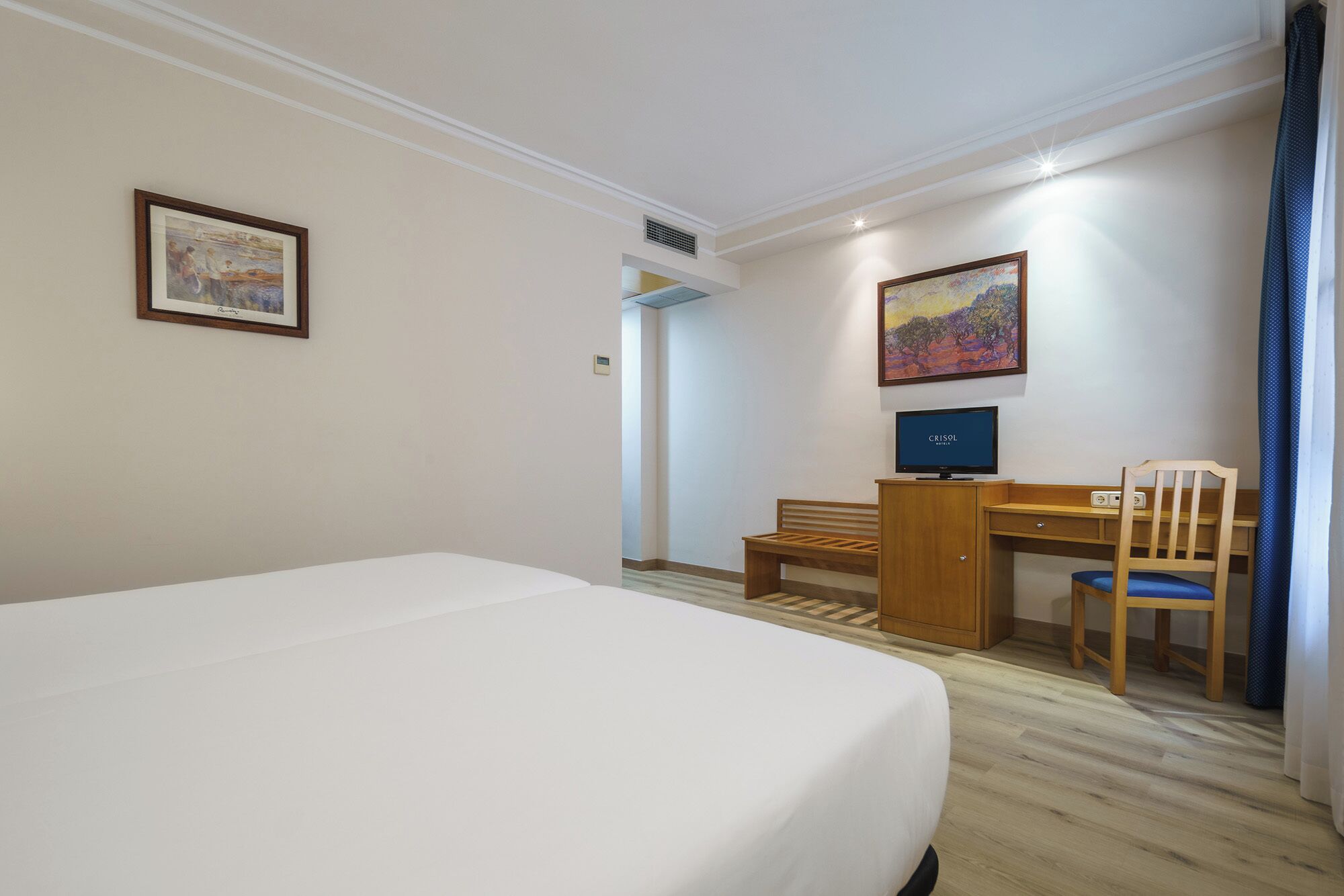 A hotel room with a bed, a desk, a chair, a television, and two paintings on the wall.