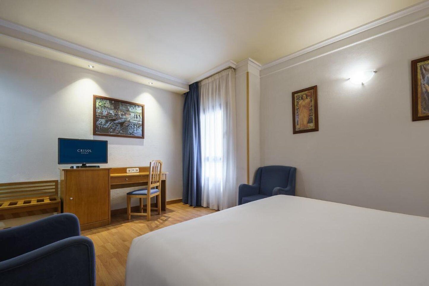Ett hotellrum med en säng, ett skrivbord med en dator, en stol och två tavlor på väggarna.