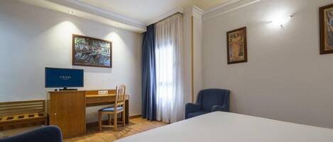 A hotel room with a bed, a desk with a computer, a chair, and two paintings on the walls.