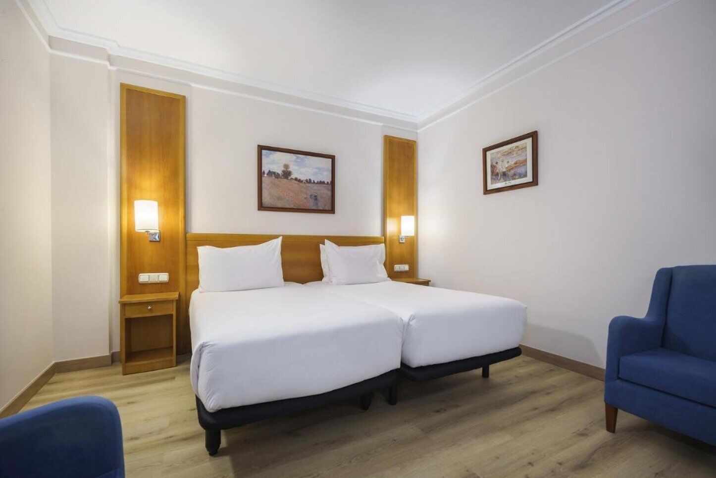 A hotel room with a double bed, two bedside tables, a blue armchair, and two wall-mounted lamps.