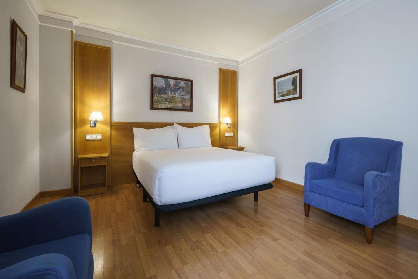 Habitación de hotel con una cama, dos lámparas de noche, un sillón azul y dos cuadros en las paredes.