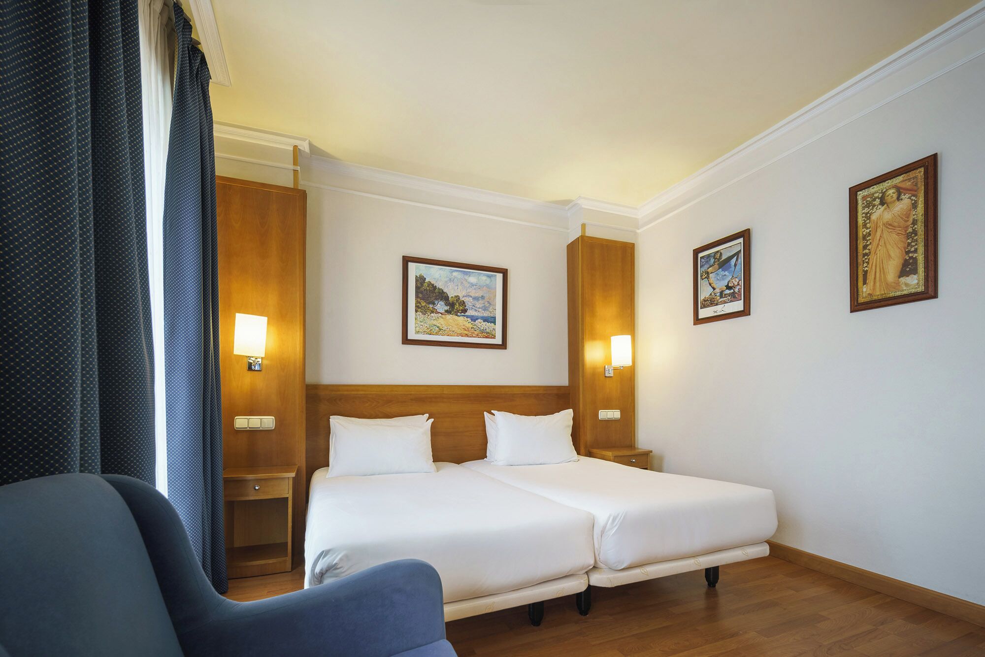 Une chambre d’hôtel avec deux lits, une tête de lit en bois, une table de chevet, une chaise et deux lampes fixées au mur.