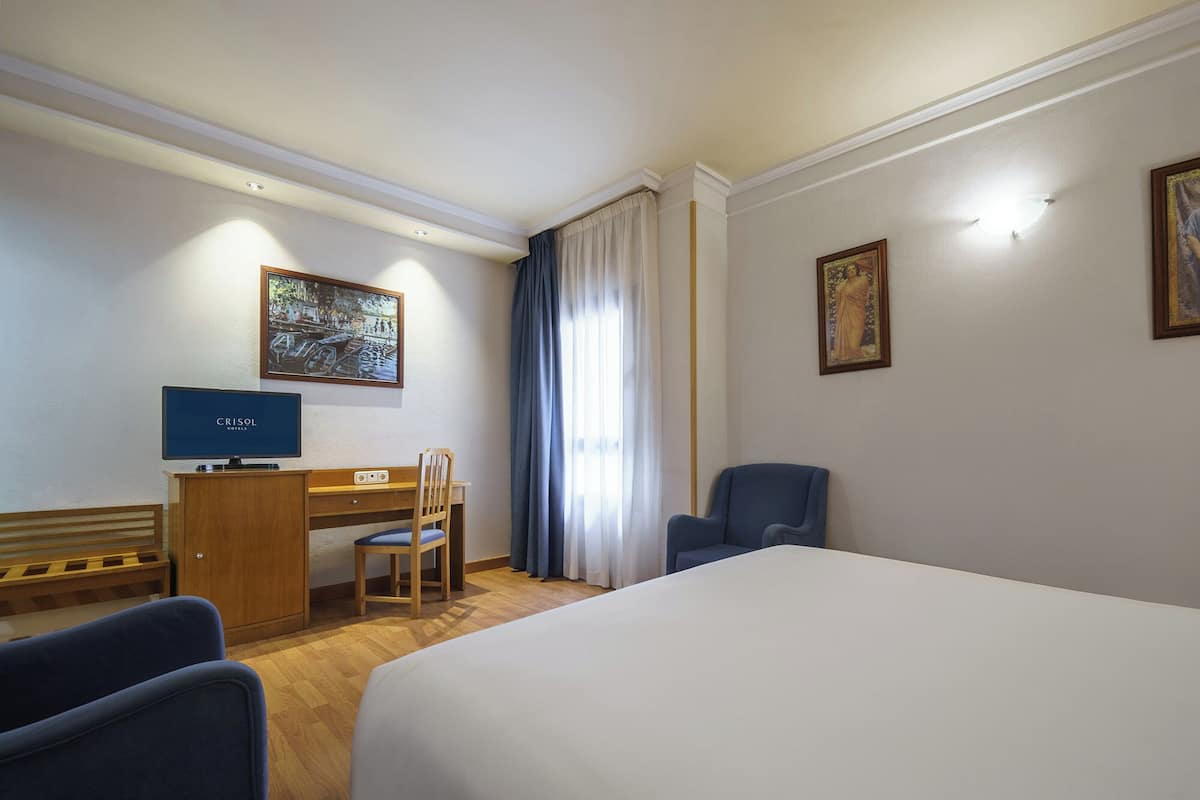A hotel room with a bed, a desk with a computer, a chair, a blue armchair, and two paintings on the wall.