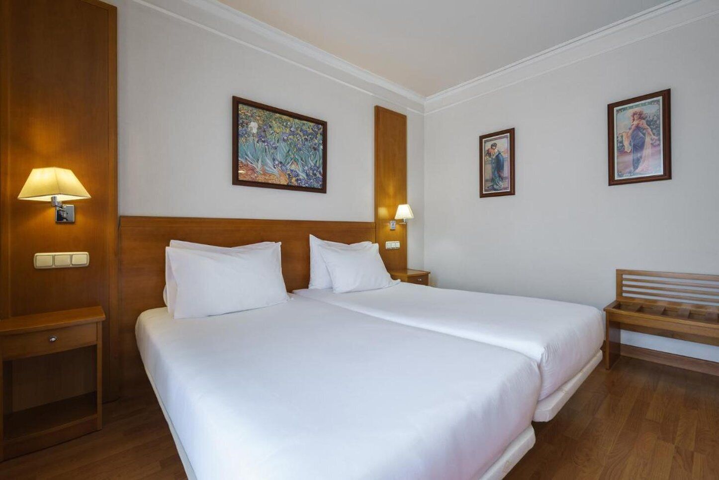 Habitación de hotel con dos camas, cabeceros de madera y un banco. Hay lámparas de pared y cuadros enmarcados.