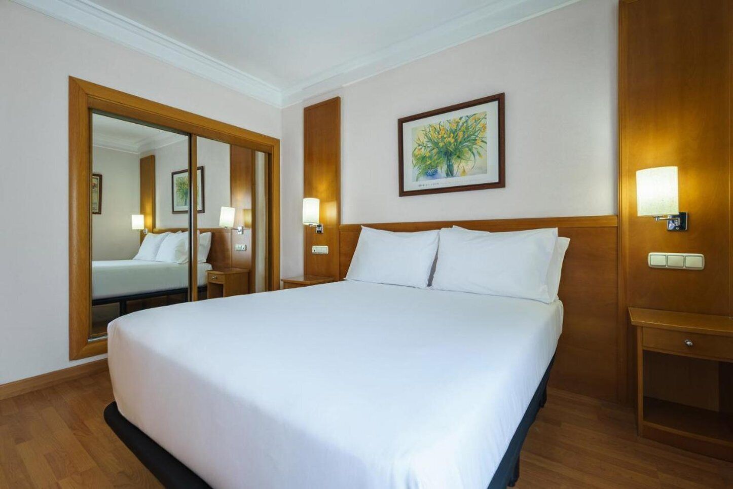 A hotel room with a large bed, a wooden headboard, a nightstand with a lamp, a mirror, and a framed picture on the wall.