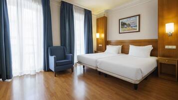A hotel room with two beds, a chair, wooden flooring, and a painting on the wall.