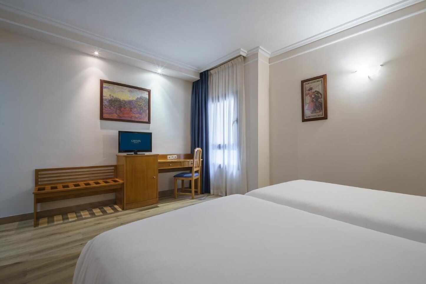 A hotel room with a bed, a desk with a computer, a chair, and two paintings on the walls.
