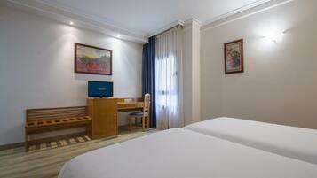 A hotel room with a bed, a desk with a computer, a chair, and two paintings on the walls.