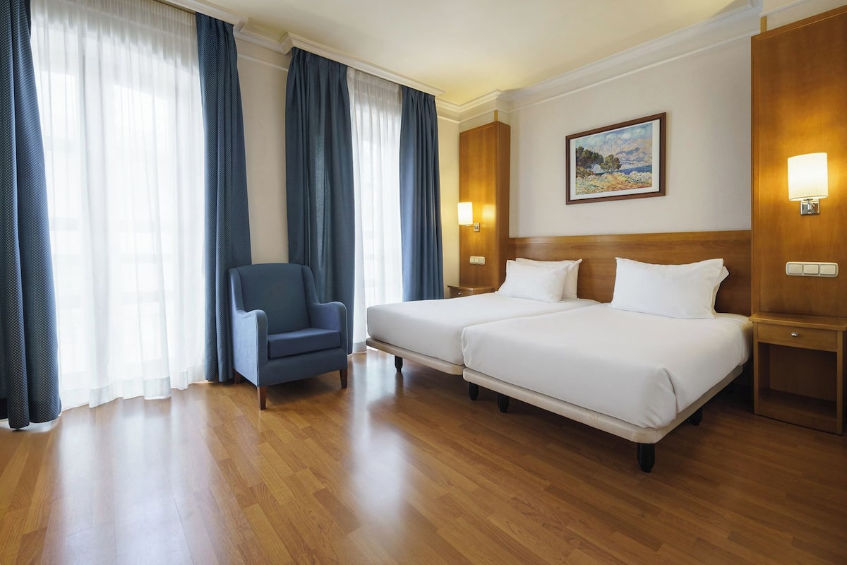A hotel room with two beds, a chair, wooden flooring, and a painting on the wall.