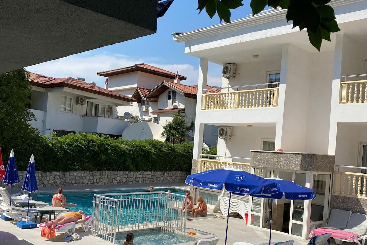 A pool area with lounge chairs and umbrellas, a swimming pool, and a building with balconies.
