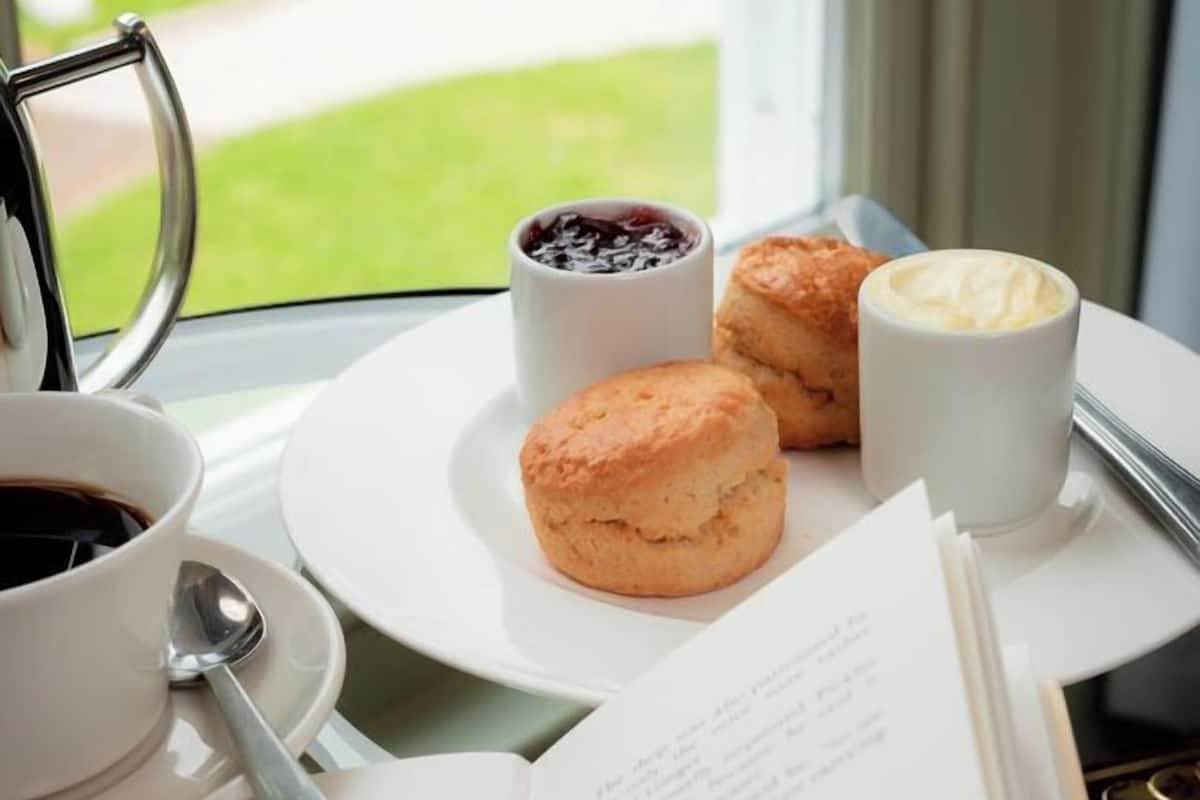 Ein Tisch mit einer Tasse Kaffee, zwei Scones, einer Marmeladetasse und einem Buch.