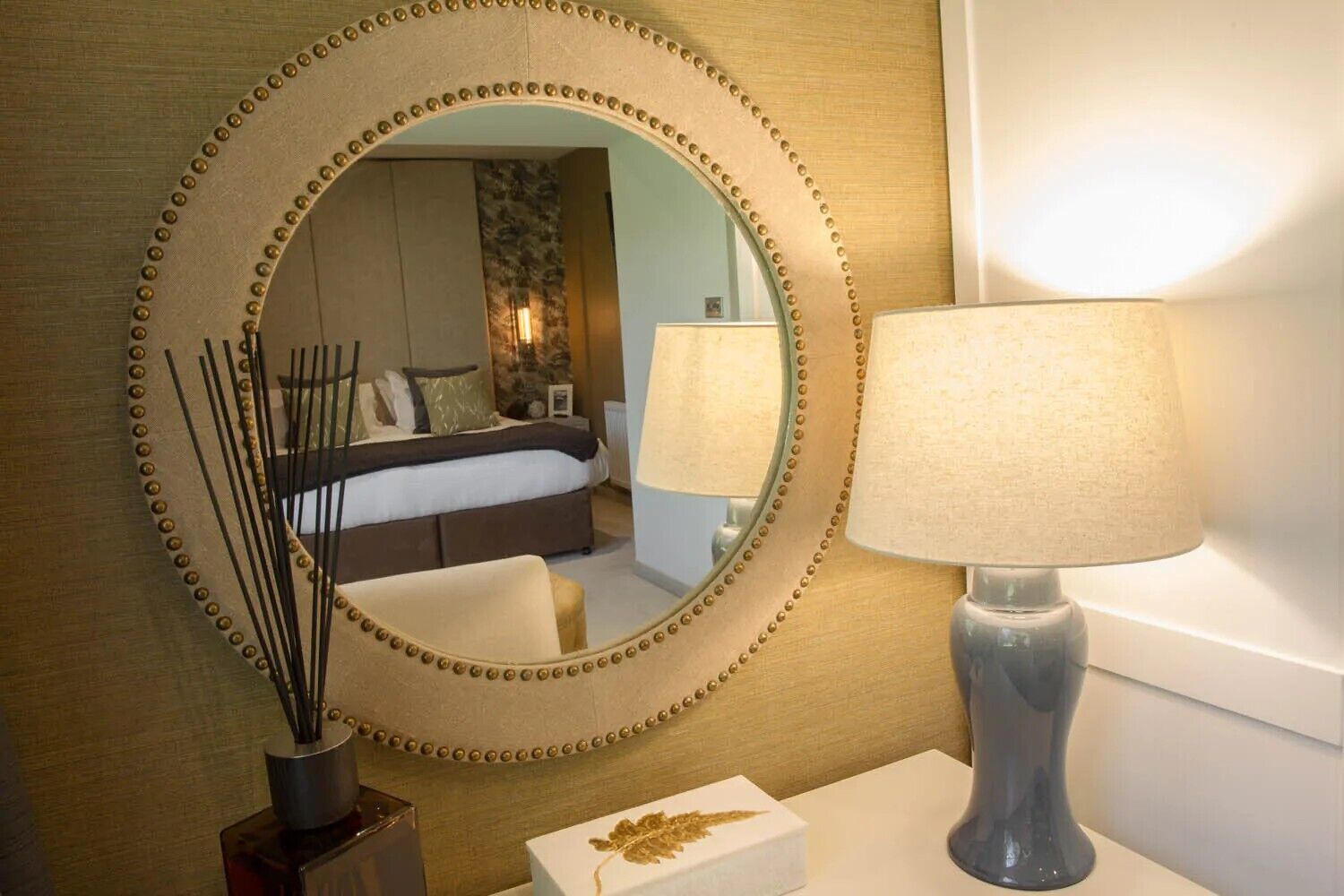 Una habitación de hotel con un espejo grande, una lámpara de noche, una mesita con un florero y una cama con cabecera.