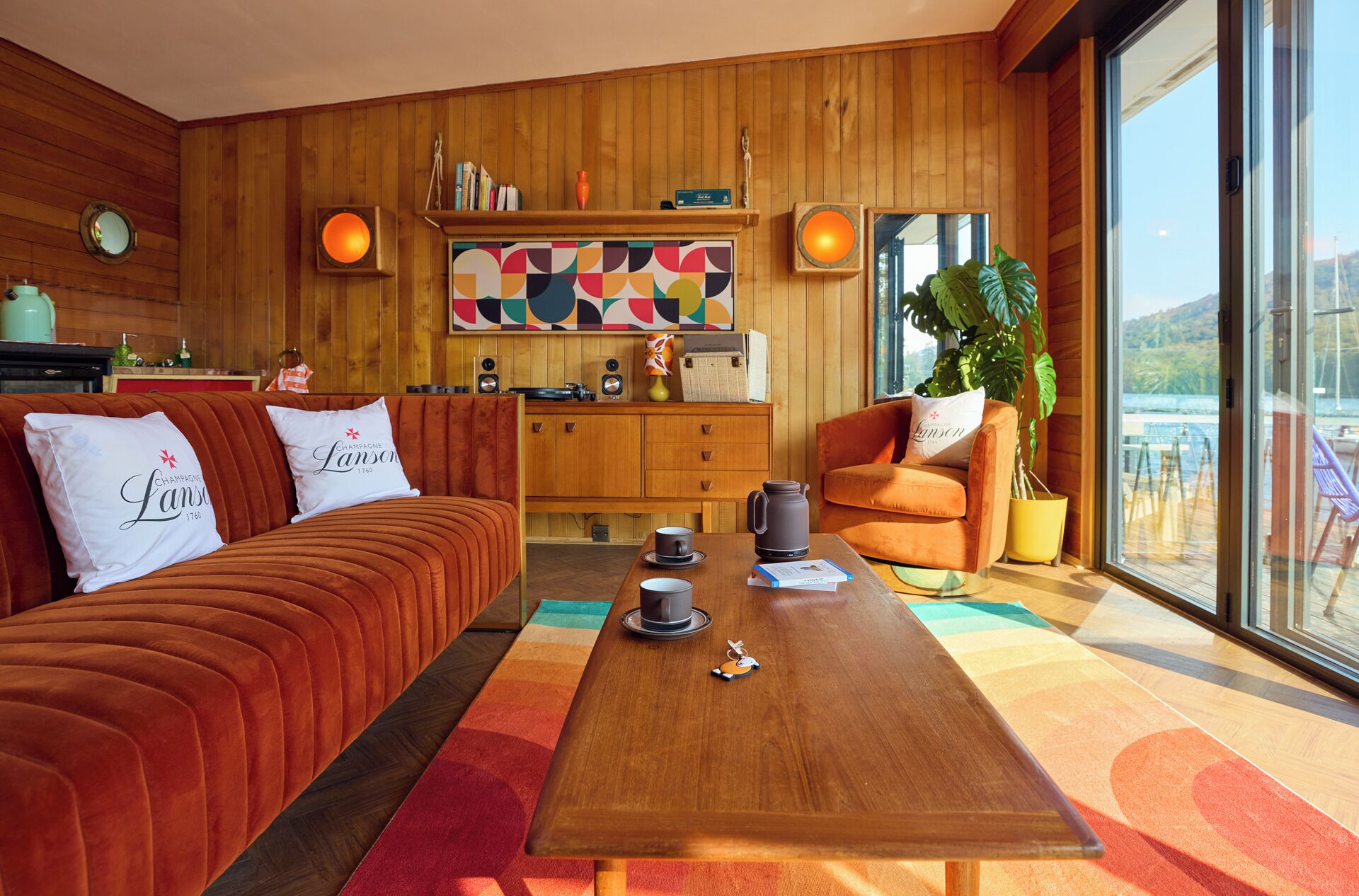 A cozy room with a wooden interior, a sofa with branded pillows, a coffee table with a teapot, and a view of a body of water through large windows.