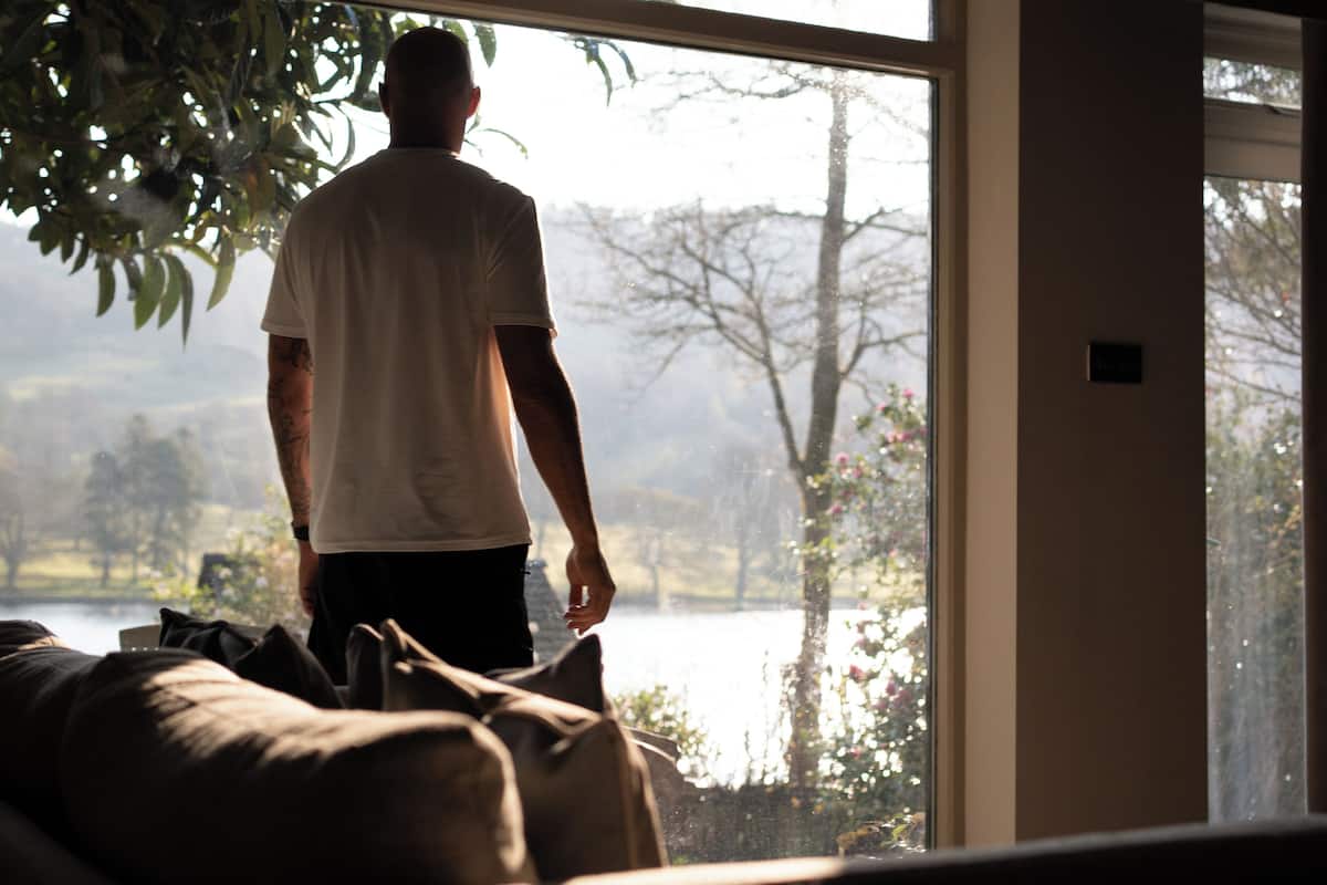 Eine Person steht an einem Fenster mit Blick auf einen See und Bäume.