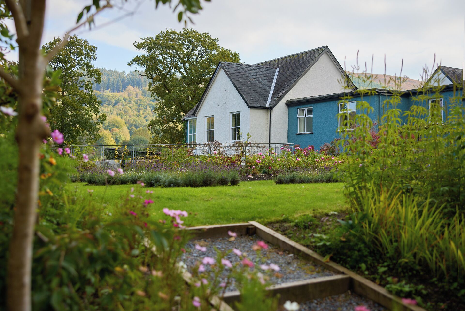 A two-story house with a white and blue exterior, surrounded by a well-maintained garden with various flowers and a gravel path.