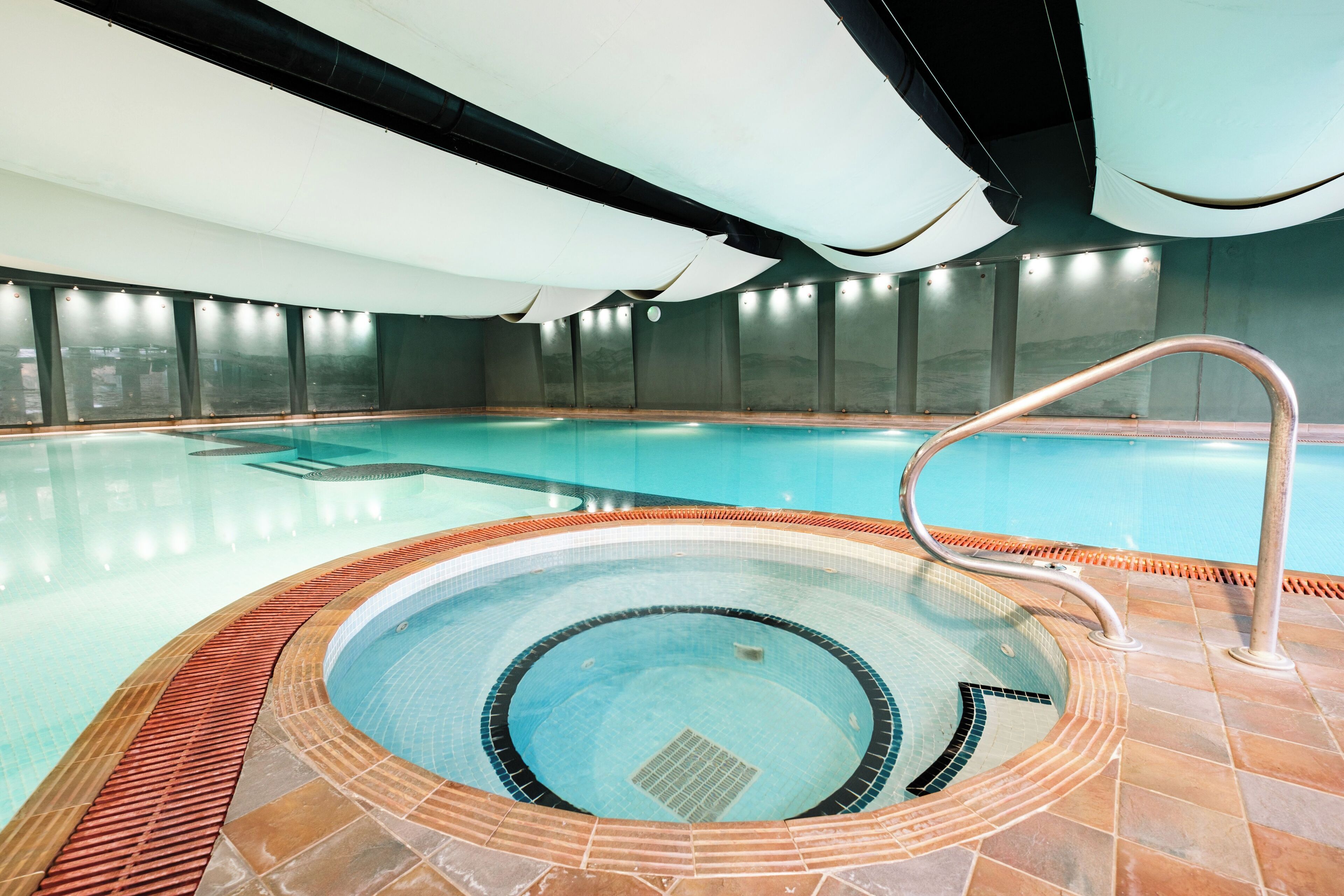 Una amplia piscina interior con un jacuzzi en el centro, rodeada de paredes y suelo con azulejos.
