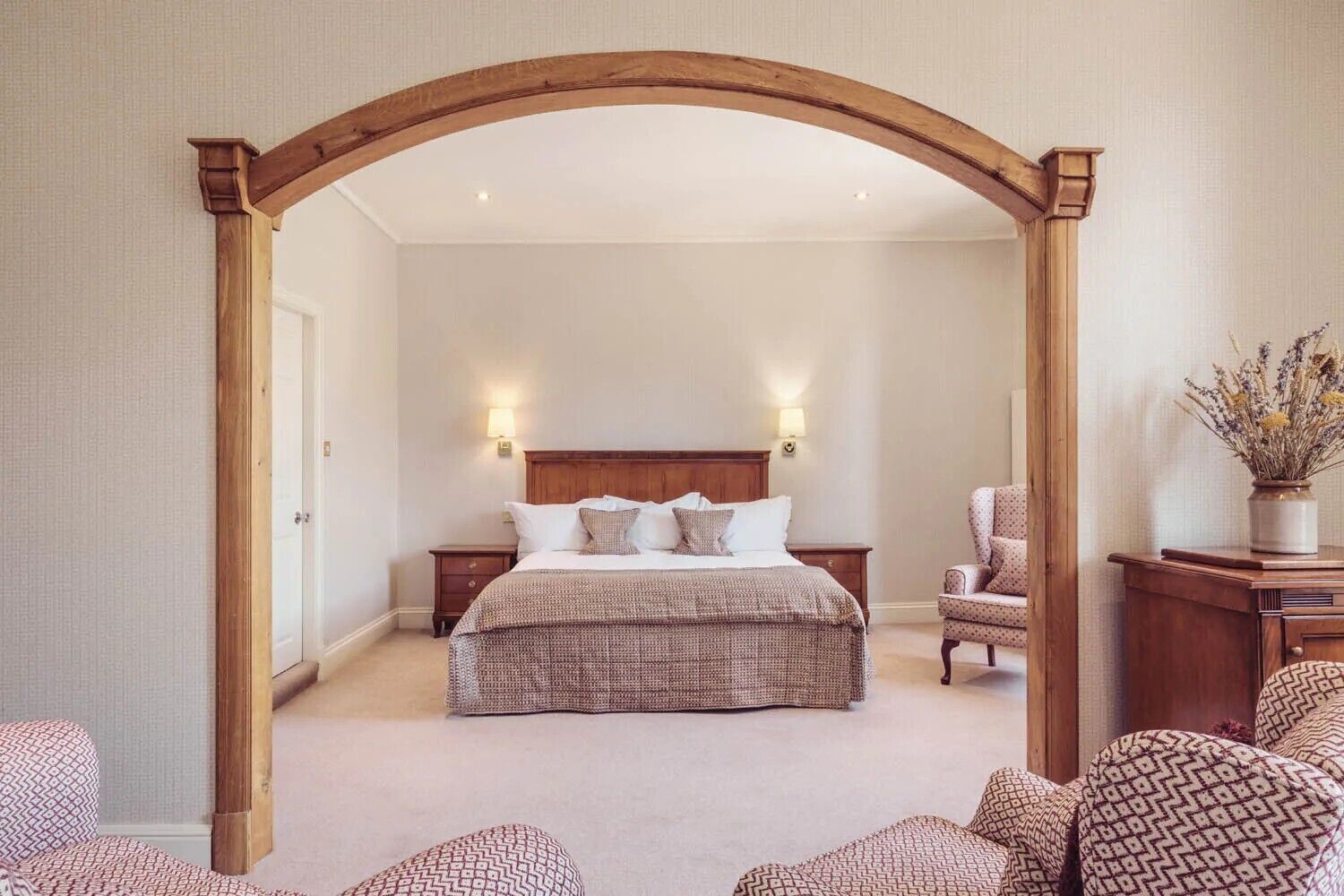 Une chambre à coucher avec un grand lit, deux tables de chevet, une porte en arc en bois et une chaise agrémentée d’un vase de fleurs.