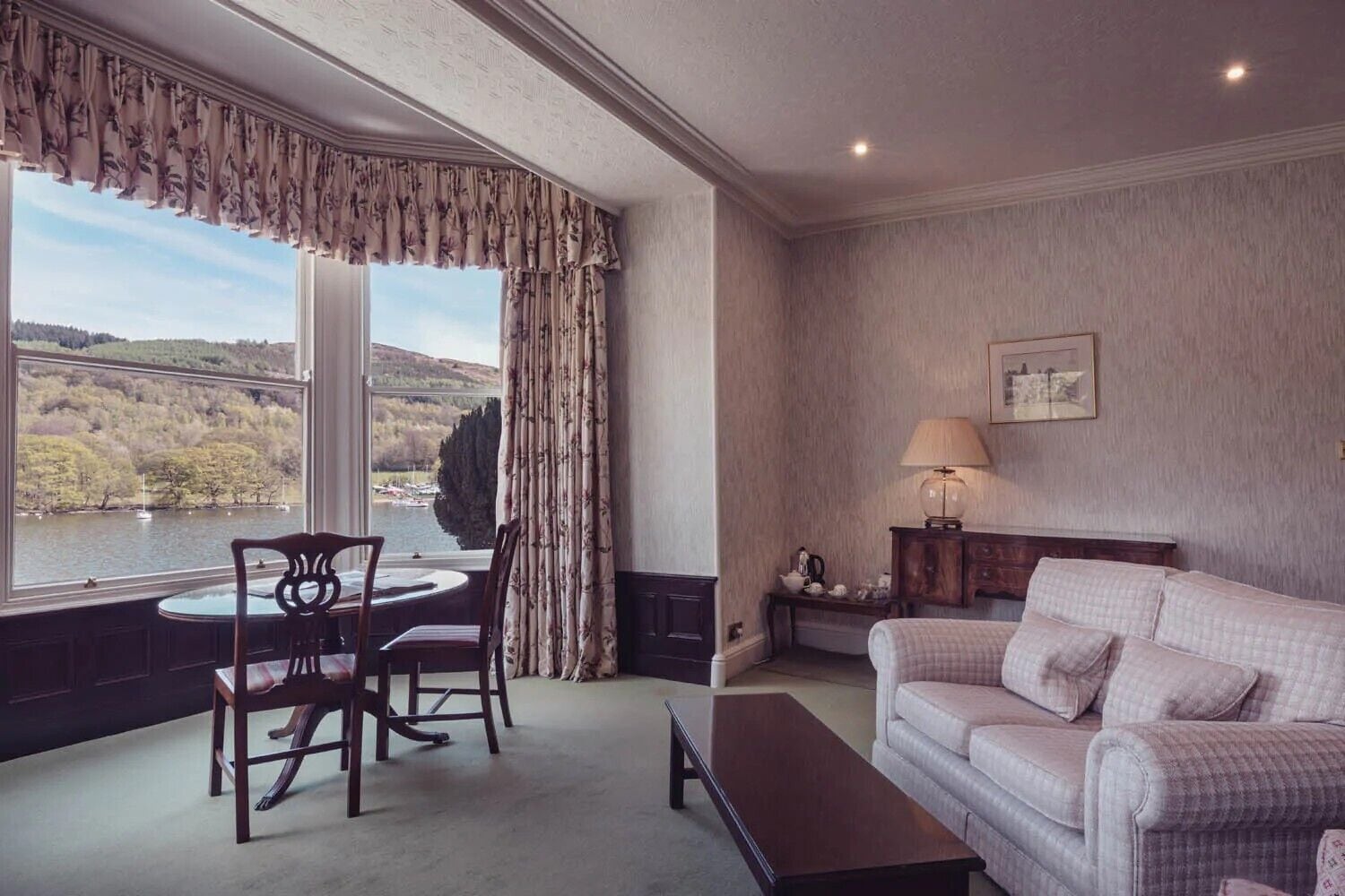 A room with a view of a lake and hills, a dining table with chairs, a sofa, and a lamp on a side table.