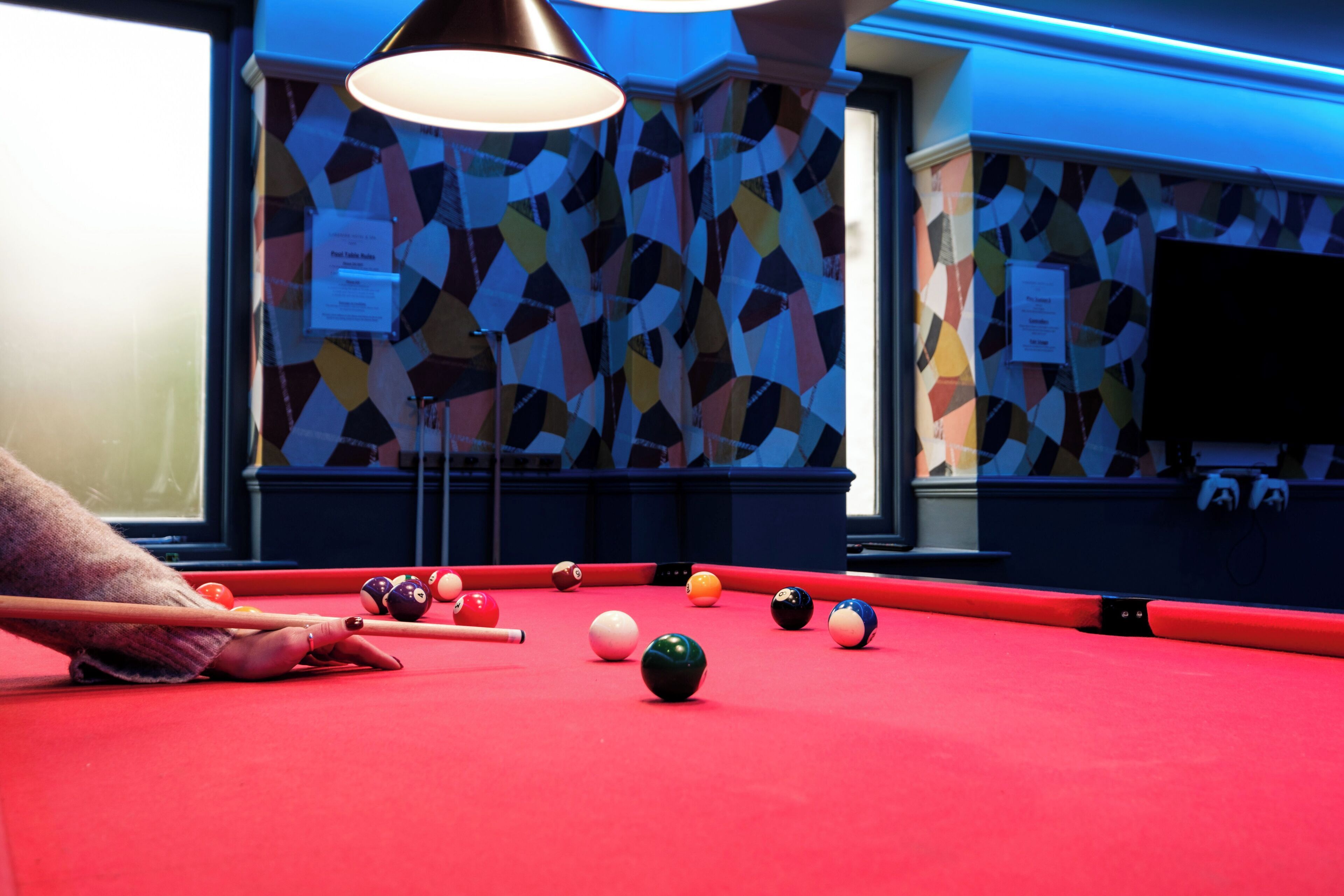 A pool table with balls and a cue stick, a patterned wall, and a television in the background.