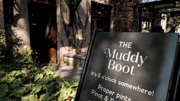 A sign for "The Muddy Boot" with text indicating it's 5 o'clock somewhere, offering proper pints, Pinot & Prosecco, and snacks.