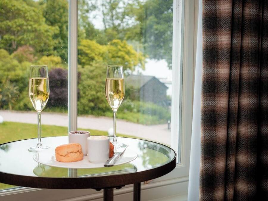 A table with two glasses of white wine, a biscuit, and a small container, set against a window with a view of greenery and a building.