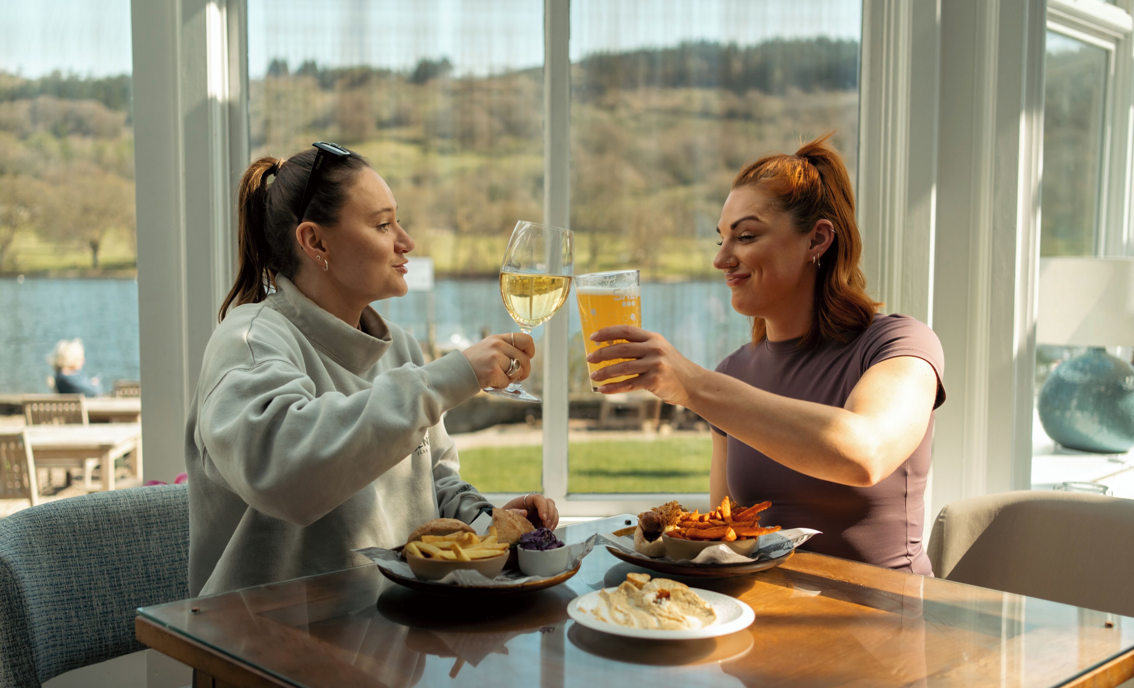 Two people toasting with drinks at a table with food and a scenic view of a lake.