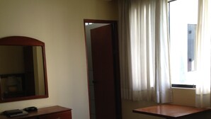 A hotel room with a bed, a desk with a chair, a mirror, and a window with curtains. - Hotel Regency (Lima)